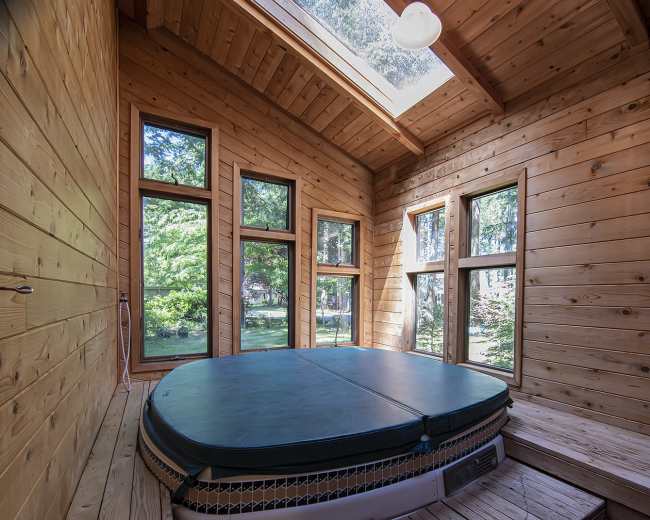 A wooden room features a circular hot tub surrounded by large windows that overlook greenery outside.