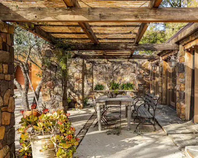 A stone patio area with a wooden pergola, featuring a dining table, chairs, and potted plants.