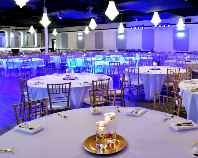 The image shows a well-decorated event space with round tables set for a banquet, illuminated by blue lighting and chandeliers.