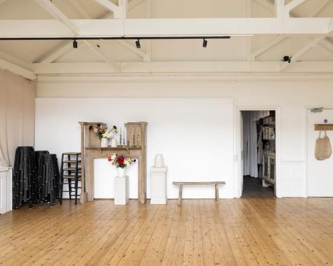 The image shows a spacious, bright room with wooden flooring, featuring a few chairs stacked against one wall, decorative shelves, a flower arrangement, a bench, and a doorway leading to another area.