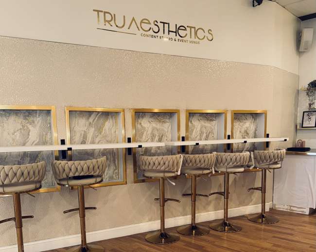 The image shows a modern interior space featuring a row of stylish bar stools arranged in front of elegant wall art, with a light-colored backdrop and a sign that reads "TRUEsthetics."
