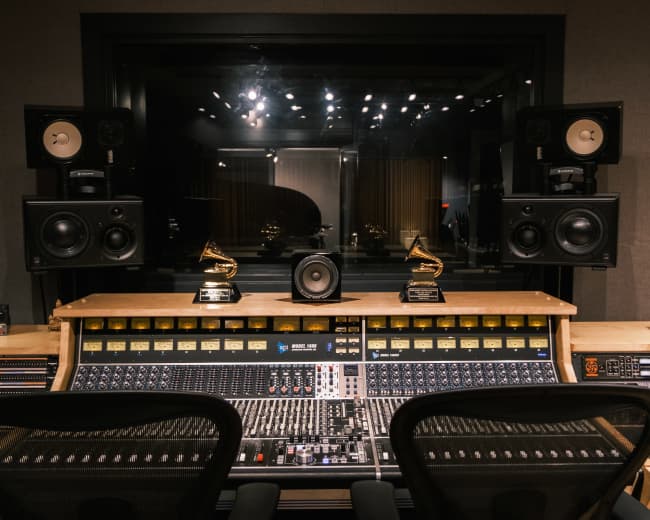 The image shows a music recording studio with a console, multiple speakers, and two Grammy trophies prominently displayed.