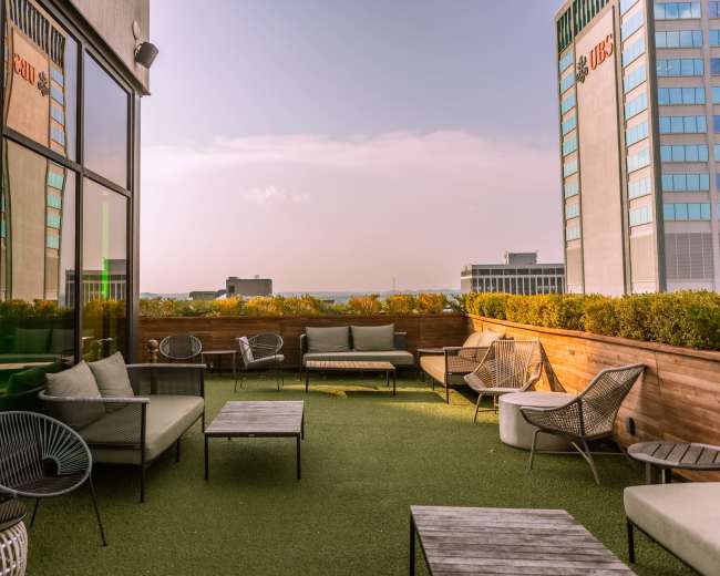 A rooftop lounge area with modern seating and greenery, set against a city skyline.