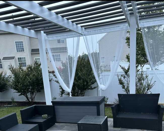 The image shows a patio area furnished with black wicker outdoor furniture, including couches, tables, and a storage chest, under a white pergola draped with sheer curtains.