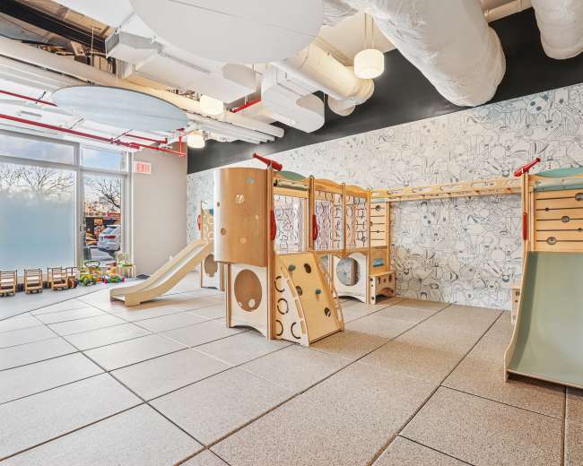 The image shows an indoor play area featuring wooden climbing structures, slides, and a textured floor in a brightly lit space.