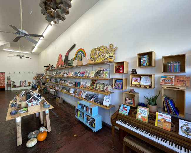 The image shows a colorful bookstore interior with wooden shelves lined with various children's books and a piano on the floor.