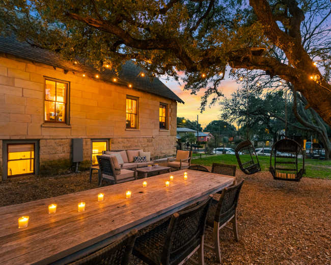 A wooden table is set with candles in an outdoor area featuring string lights and a swing, with a building and trees in the background at sunset.