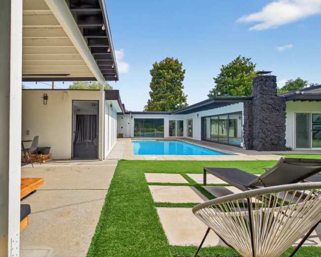 The image depicts a modern outdoor living space featuring a pool, lounge chairs, and a stone fireplace, surrounded by greenery and contemporary architecture.