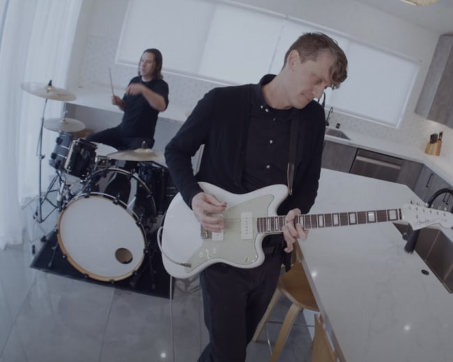 A band performs in a modern kitchen with a drummer, guitarist, and bassist.