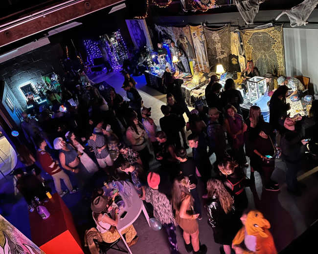 A crowded indoor venue features various decorations and people engaged in conversation, with booths set up in the background.