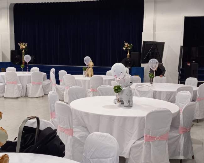 A spacious event hall features round tables dressed in white cloth with pink sashes, and a stage at the front with a dark curtain.