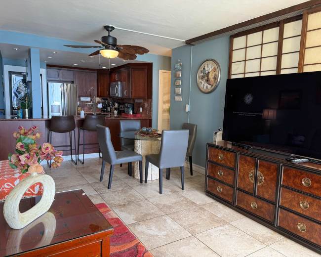 The image shows a living area featuring a dining table, kitchen in the background, a television, and various pieces of furniture.