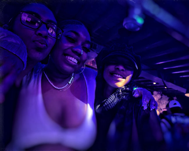 Three women pose together in a dimly lit venue illuminated by purple light.