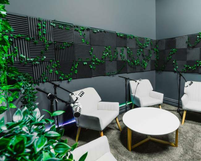 The image shows a small recording studio with three white chairs, a round table, and soundproofed walls adorned with green foliage.