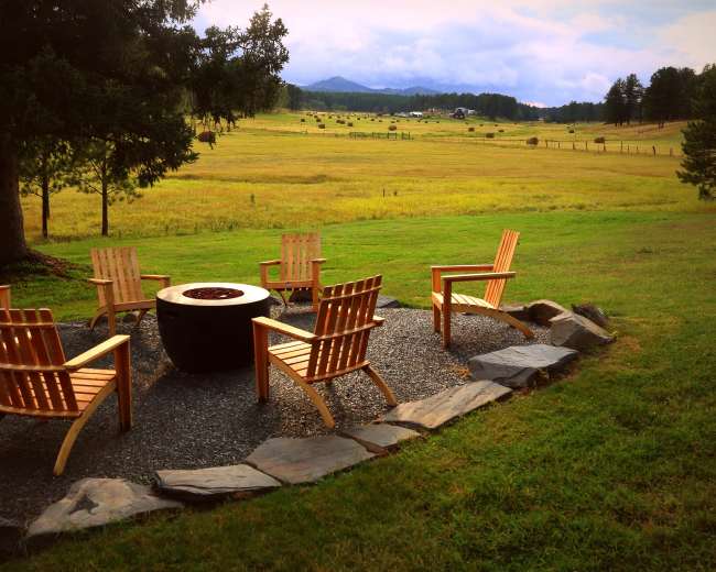 A circular arrangement of wooden chairs surrounds a stone fire pit in a grassy area with a backdrop of rolling hills.