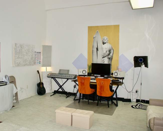 The image shows a modern studio space featuring a desk with two orange chairs, a computer setup, a keyboard, and decorative elements including a large artwork of a statue and a surfboard on the wall.