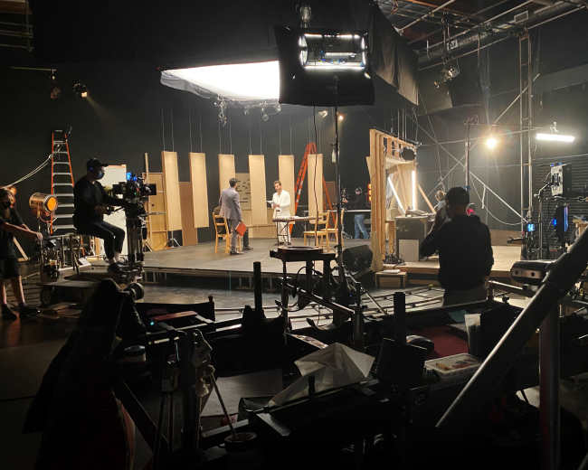 A film or television production set features crew members and equipment arranged around a central stage with lighting and backdrop elements.
