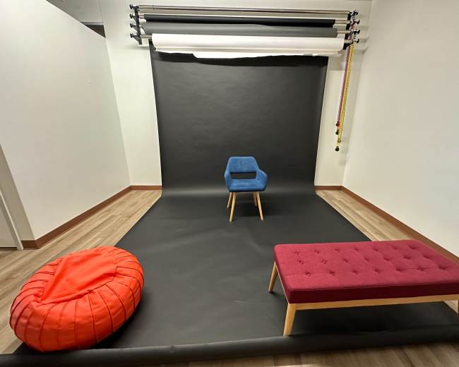A minimalist studio setup with a black backdrop, a blue chair on a wooden frame, a red bean bag, and a purple bench.