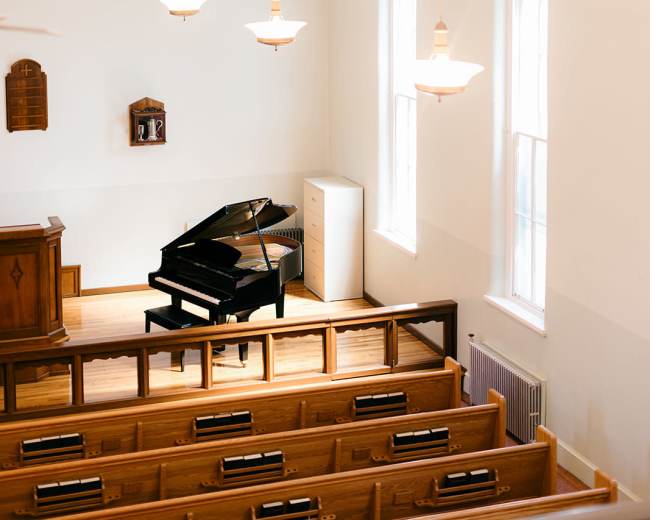 The image shows a view of an interior of a church with a grand piano prominently placed in front of a wooden podium, flanked by rows of wooden pews and large windows.