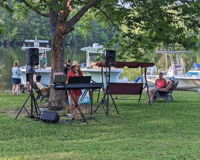 A musician plays keyboards near the water while people relax on the shore and boats are docked nearby.
