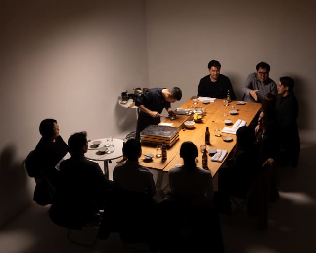 A group of eight people gathers around a large wooden table, while one person prepares food on a grill in the center.