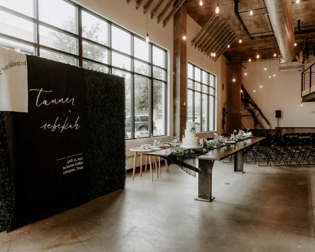 The image shows an event space with large windows, exposed ceiling beams, and a decorated area featuring a dark green backdrop and a long table set for a gathering.