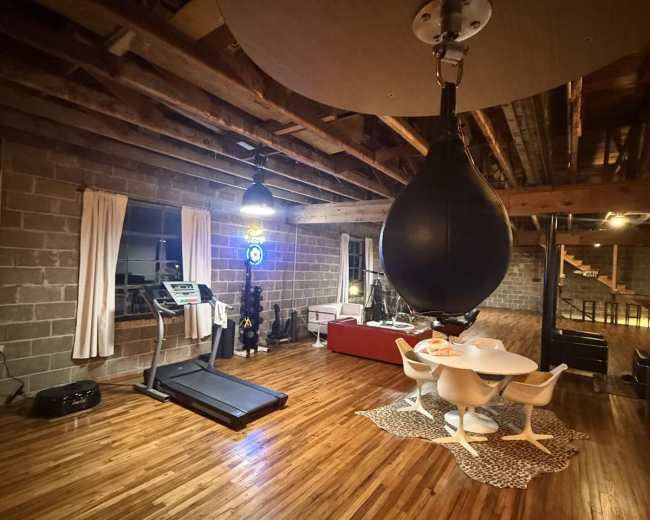 The image shows a spacious room with wooden floors featuring a treadmill, a red couch, a dining table with white chairs, and a hanging punching bag.