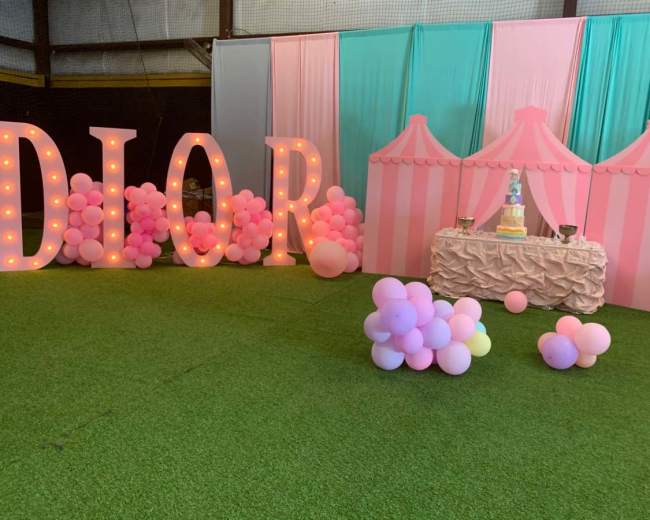 The scene features large illuminated letters spelling "DIOR," pastel-colored tents, and a decorated table with a cake, all arranged on a grass-like surface.