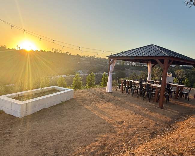 A wooden gazebo with a large table and chairs is positioned on a hillside overlooking a sunset.