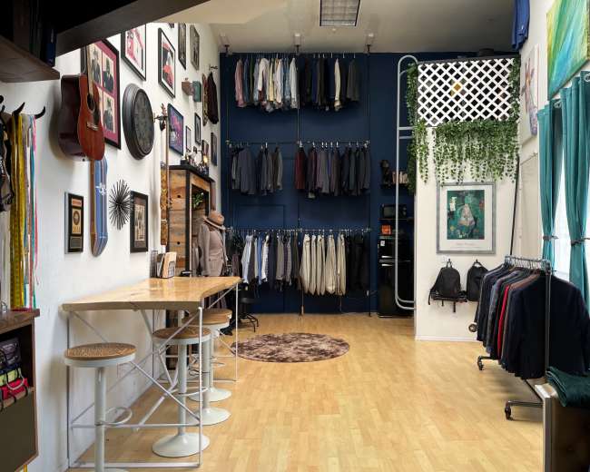 A clothing store interior with a variety of suits hanging on the wall, a round rug on the floor, and a table with stools.