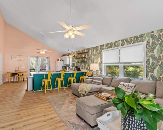 The image shows a bright, open living space with a sectional sofa, an island bar with yellow stools, and a kitchen in the background adorned with tropical-themed wallpaper.
