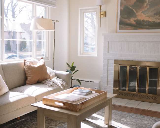 The image shows a bright living room with a beige sofa, a wooden coffee table, and a fireplace, illuminated by natural light from large windows.