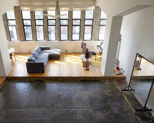 The image shows a spacious loft-style room with large windows, featuring a gray couch, a coffee table, and a small music setup in the corner.