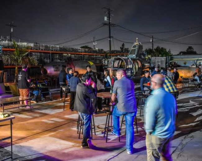 A lively outdoor gathering is taking place at night, featuring people socializing around tables amid industrial surroundings.