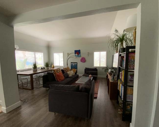 The image shows a bright living space with a seating area featuring dark couches, a dining table, bookshelves, and various plants in a modern interior design.