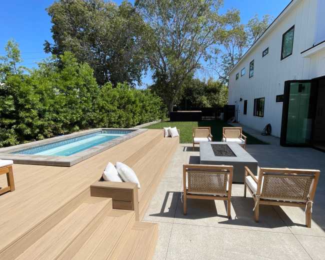 A backyard features a wooden deck with a plunge pool, a concrete table surrounded by chairs, and a green lawn bordered by shrubs and trees.