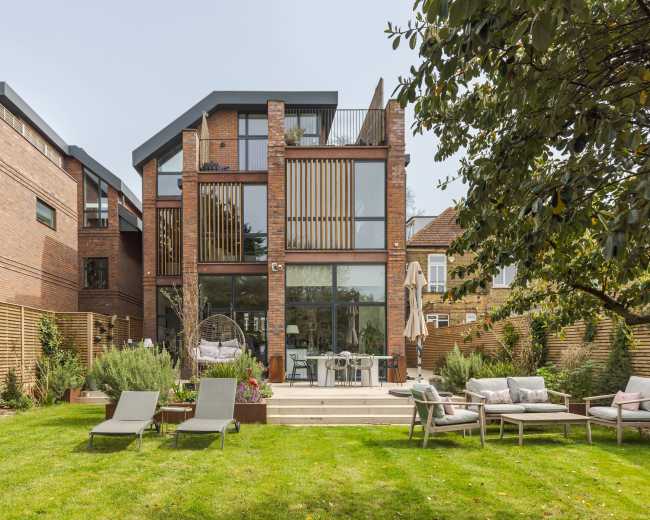 The image shows a modern brick house with large glass windows and a landscaped backyard featuring lounge chairs and outdoor furniture.