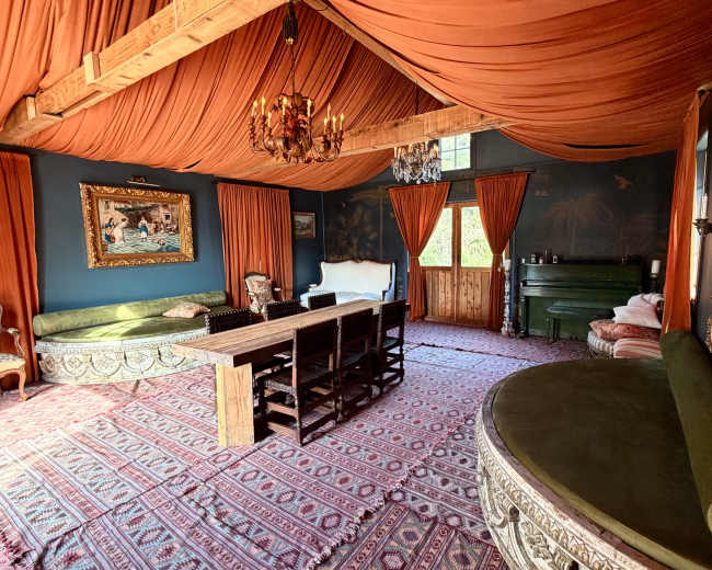 The image shows a room with orange draped fabric covering the ceiling, a long wooden dining table surrounded by black chairs, and a green piano against a wall adorned with a painting.