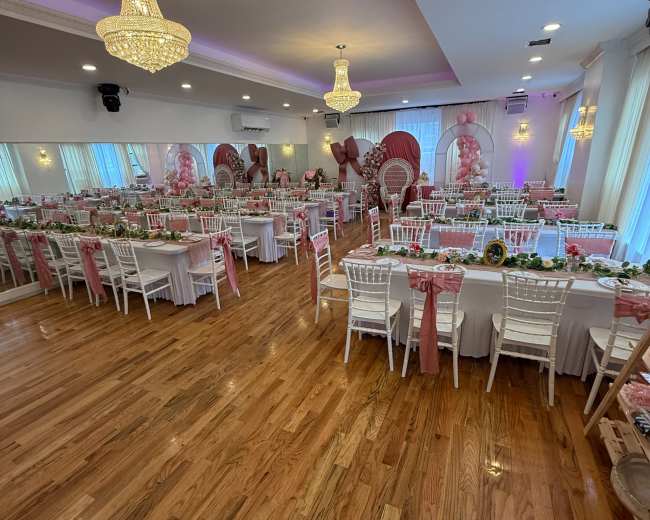 The image shows a decorated banquet hall with white tables set for an event, featuring pink accents, floral centerpieces, and elegant chandeliers.