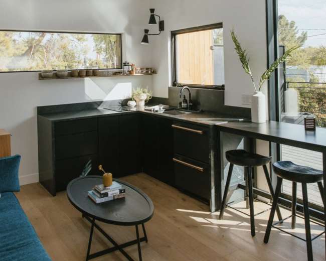 The image shows a modern kitchen nook with black cabinetry, a small dining table, and large windows allowing natural light to fill the space.