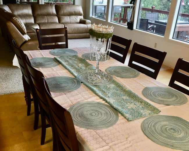 A dining table is set with a light tablecloth and decorative placemats, surrounded by wooden chairs in a brightly lit room with large windows.