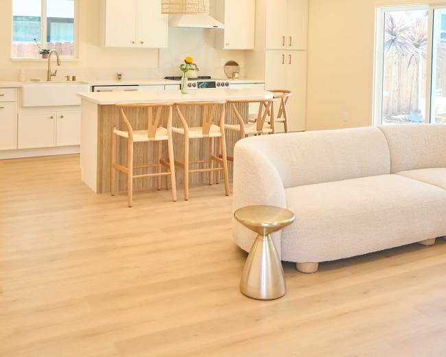 The image features a modern, open-concept kitchen and living area with a light color palette, wooden accents, and minimalistic furniture.