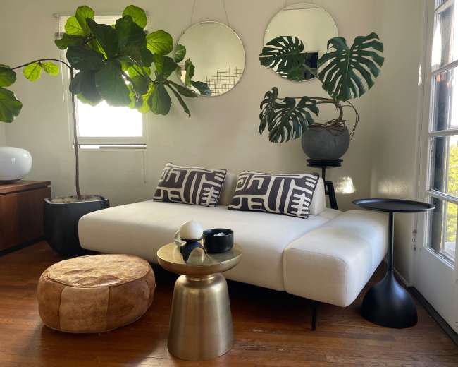 The image features a modern living room with a white couch, patterned cushions, a round brass coffee table, a black side table, and large potted plants beside mirrors on the wall.