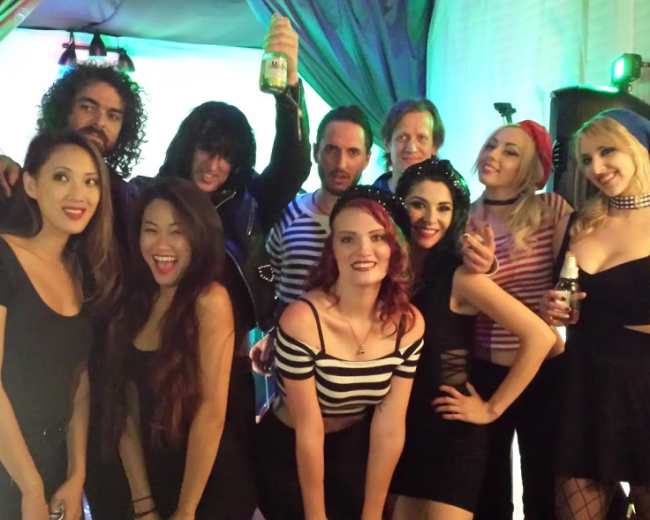 A group of ten people poses together in a brightly lit room, smiling and holding drinks.