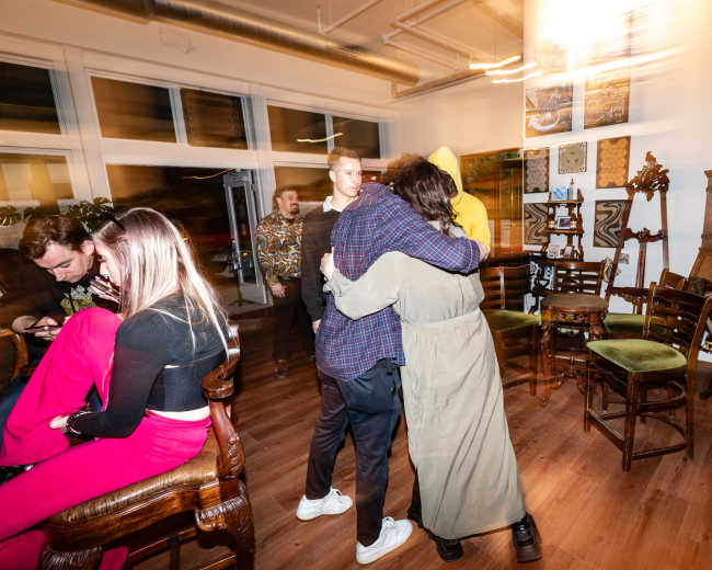 A group of people interacts in a warmly lit room filled with chairs and artwork, with two individuals embracing in the foreground.