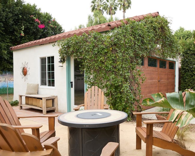 The image shows a small white cottage with a red tile roof, surrounded by lush greenery and outdoor seating made of wooden chairs arranged around a round table.