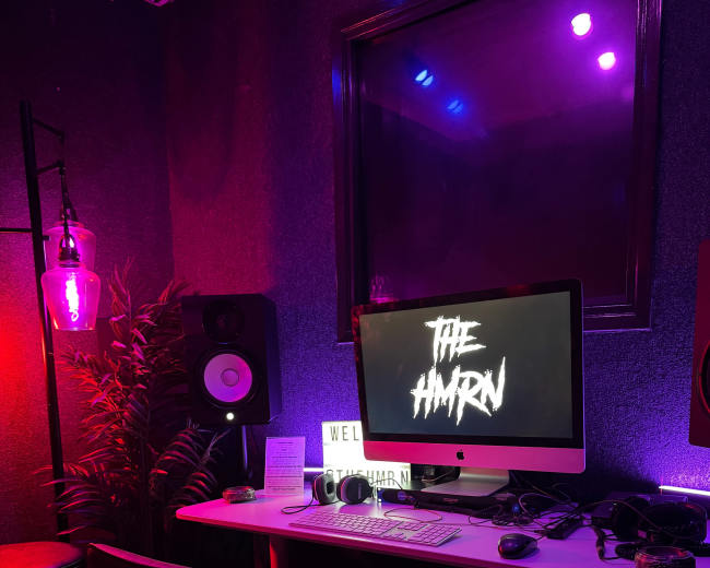 A modern computer desk is set up in a dimly lit room with colorful lighting, featuring a large monitor displaying "THE HAMRN," surrounded by audio equipment and decorative plants.