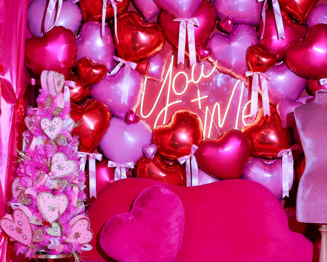 A bright pink backdrop displays a heart-themed arrangement of balloons and a neon sign reading "You + Me," alongside a decorative Christmas tree and a pink couch.
