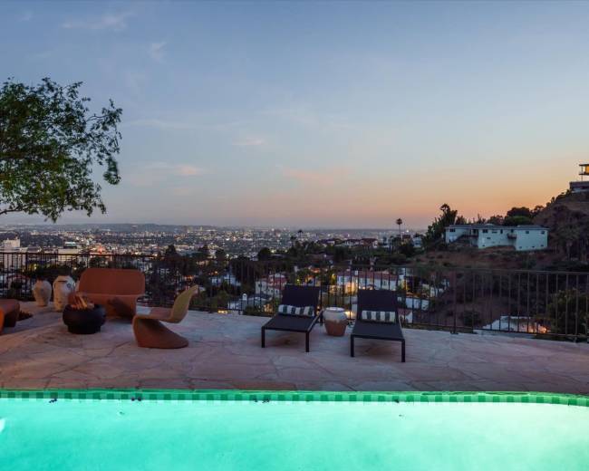 A poolside patio overlooks a sprawling city skyline at dusk, with comfortable seating arranged around a fire pit.