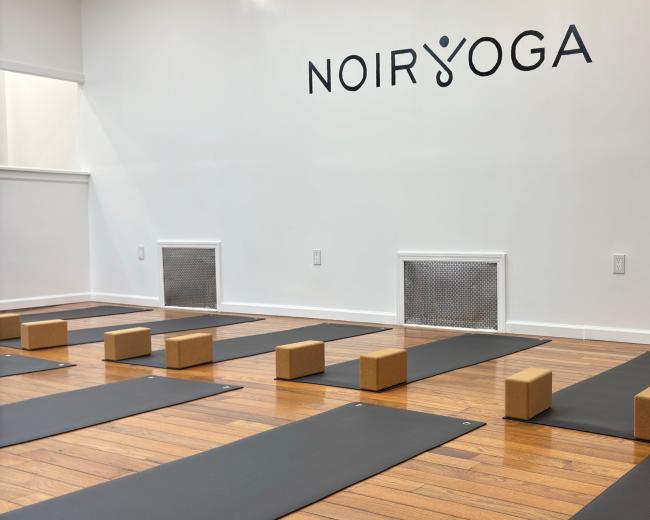 The image shows a yoga studio with several yoga mats neatly arranged on a wooden floor, and yoga blocks positioned at intervals along the mats, all against a wall with the logo "NOIR YOGA."